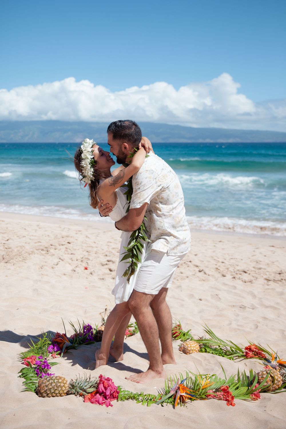 A couple shares romantic moments on a tropical beach decorated with vibrant flowers and a pineapple on the sand.