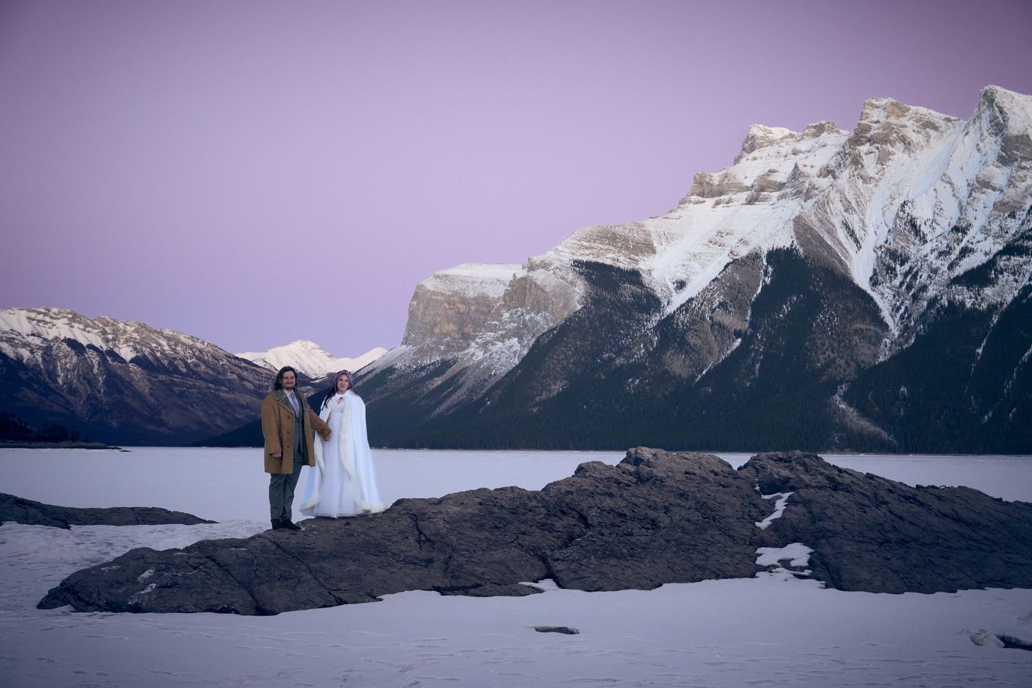 A scenic winter wedding photo shoot at Lake Minnewanka in Banff National Park with snow-capped mountains in the background.