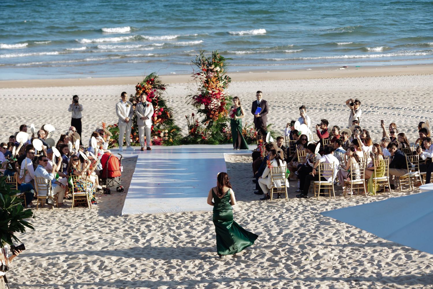 Aerial view of a colorful beach wedding ceremony with guests seated along a white aisle.