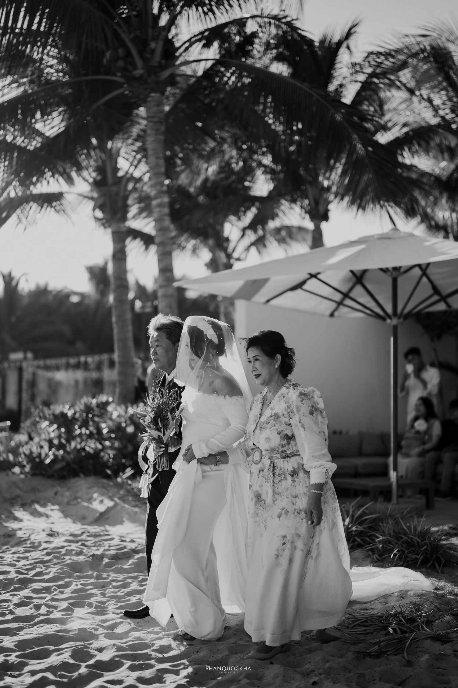 Black and white photo of a bride walking down the aisle with family on a tropical beach.
