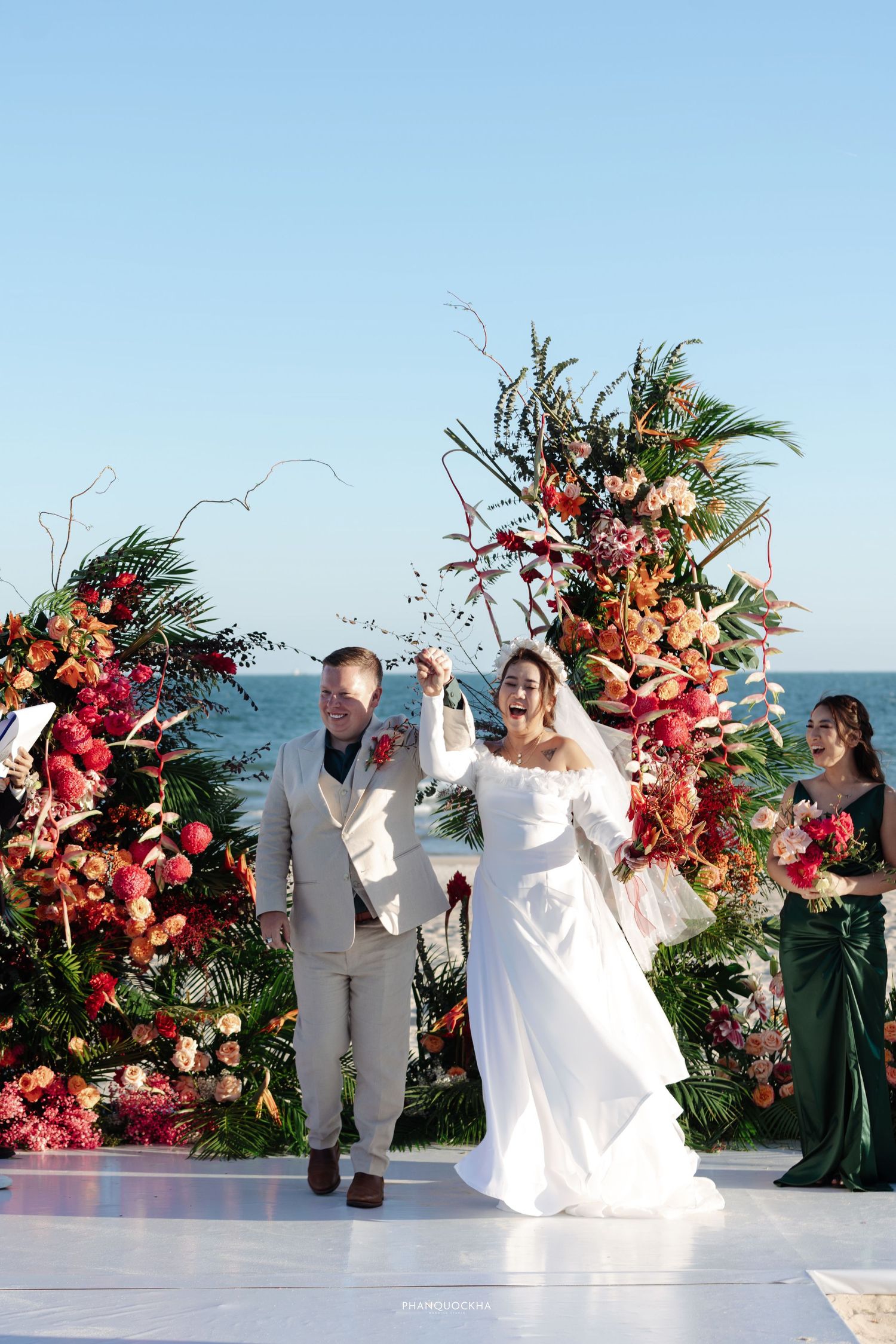 Joyful bride and groom celebrate at altar with vibrant tropical floral arch by the ocean.