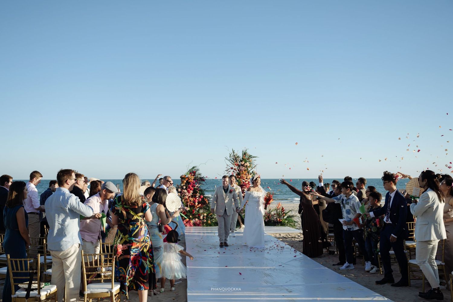 Wedding guests toss petals as bride and groom walk down the aisle at a beach ceremony.