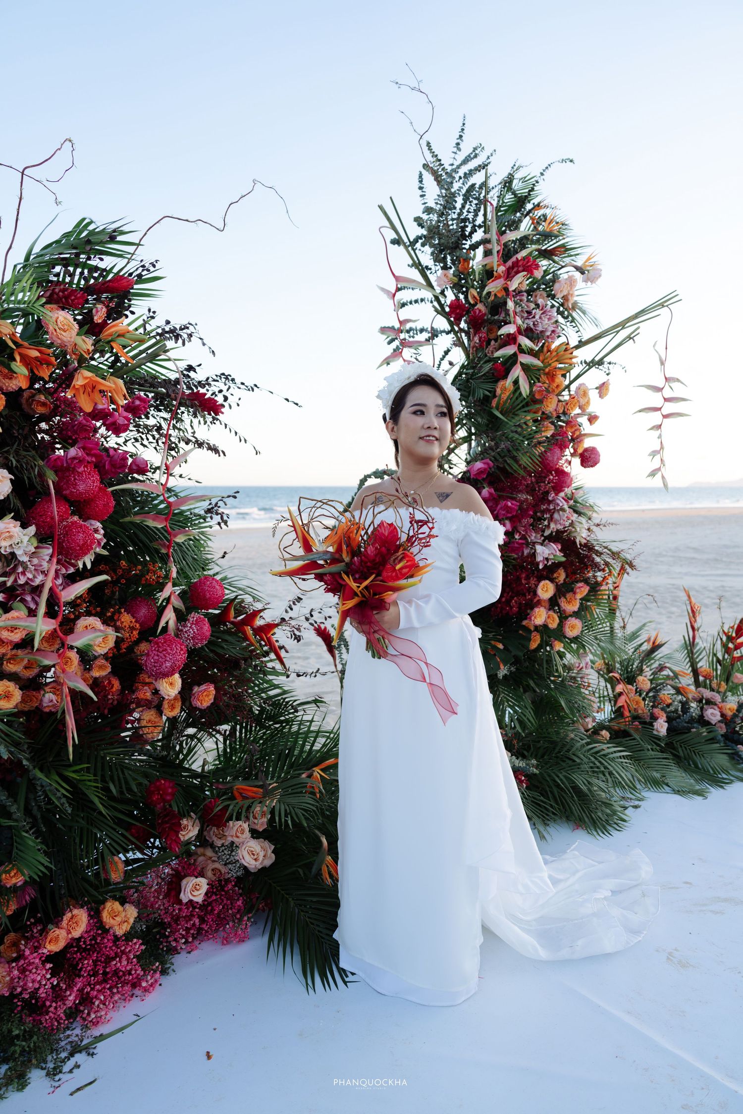Bride in white off-shoulder gown poses before a vibrant tropical floral arch on the beach.
