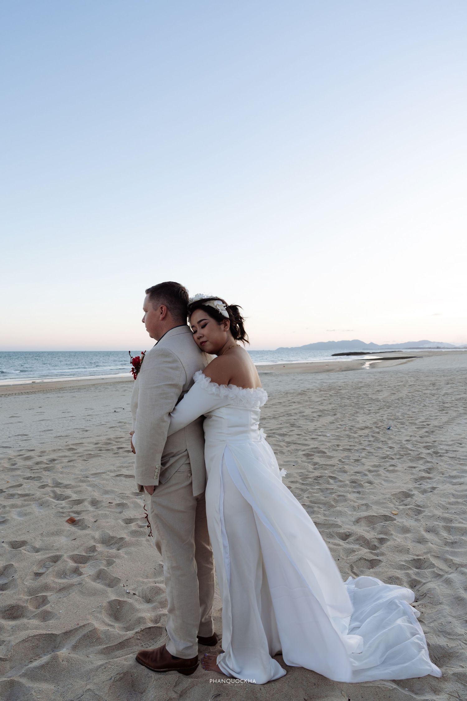 Bride embraces groom from behind on a serene sandy beach at sunset with mountains afar.