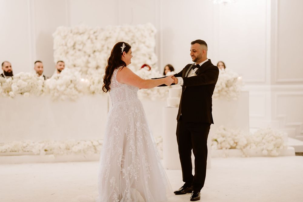 Wedding couple dancing in an elegant white reception hall decorated with floral arrangements.