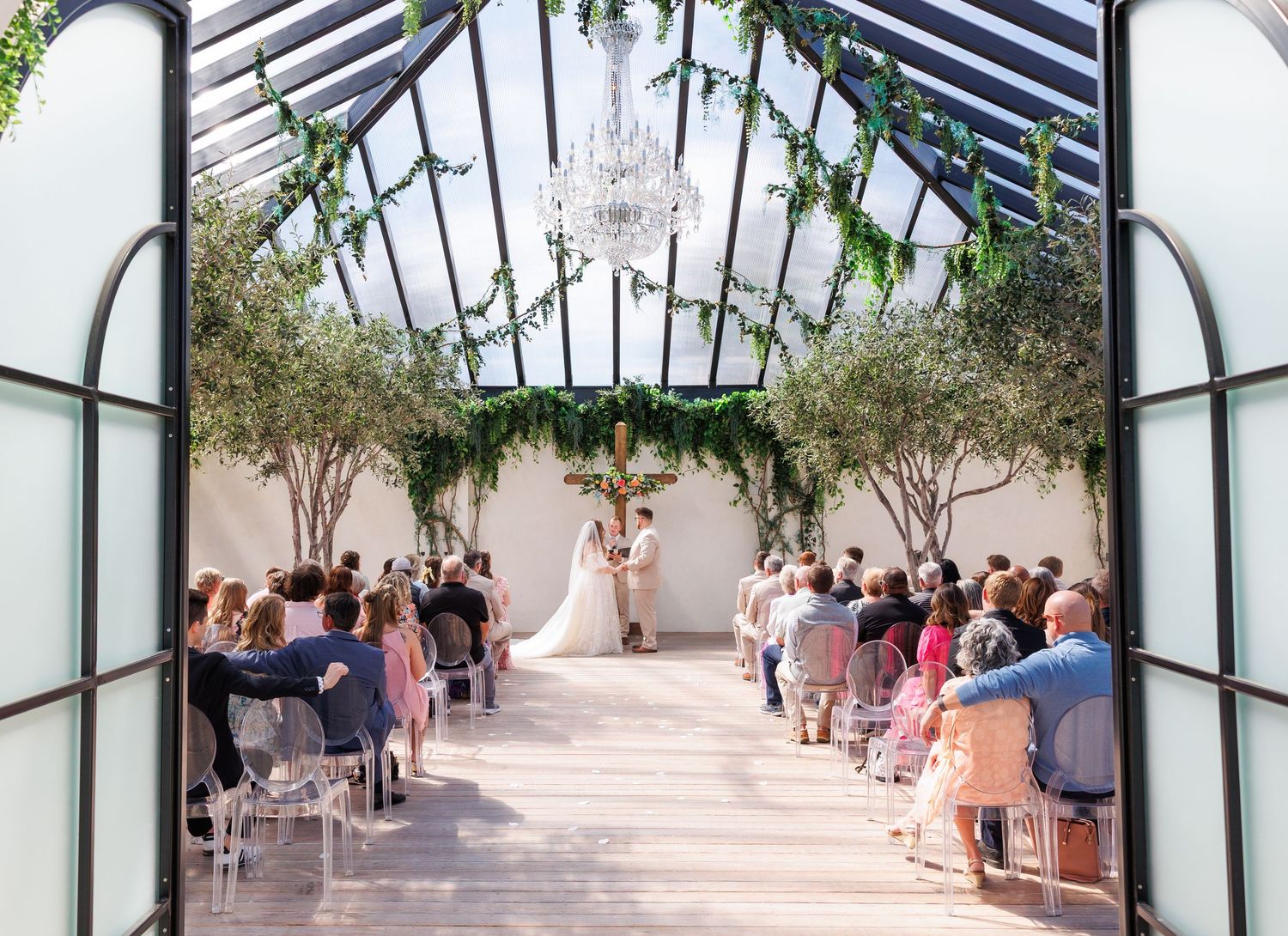 A panoramic view of guests gathering at an outdoor ceremony space with arched glass doors and garden surroundings at Greenhouse Two Rivers.