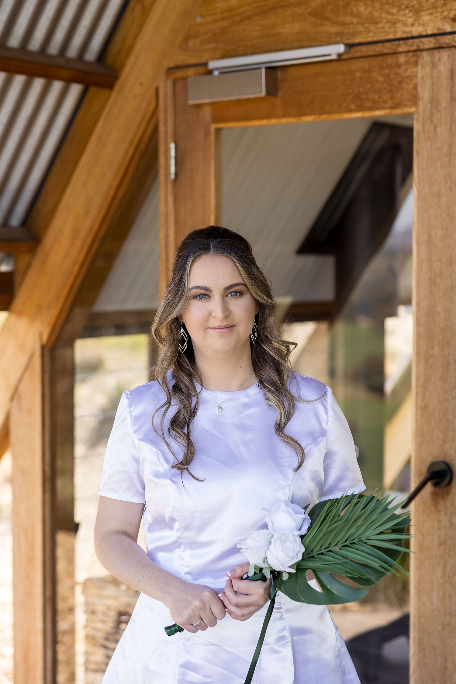 Woman in white dress poses outdoors under rustic wooden beams holding a large tropical leaf.