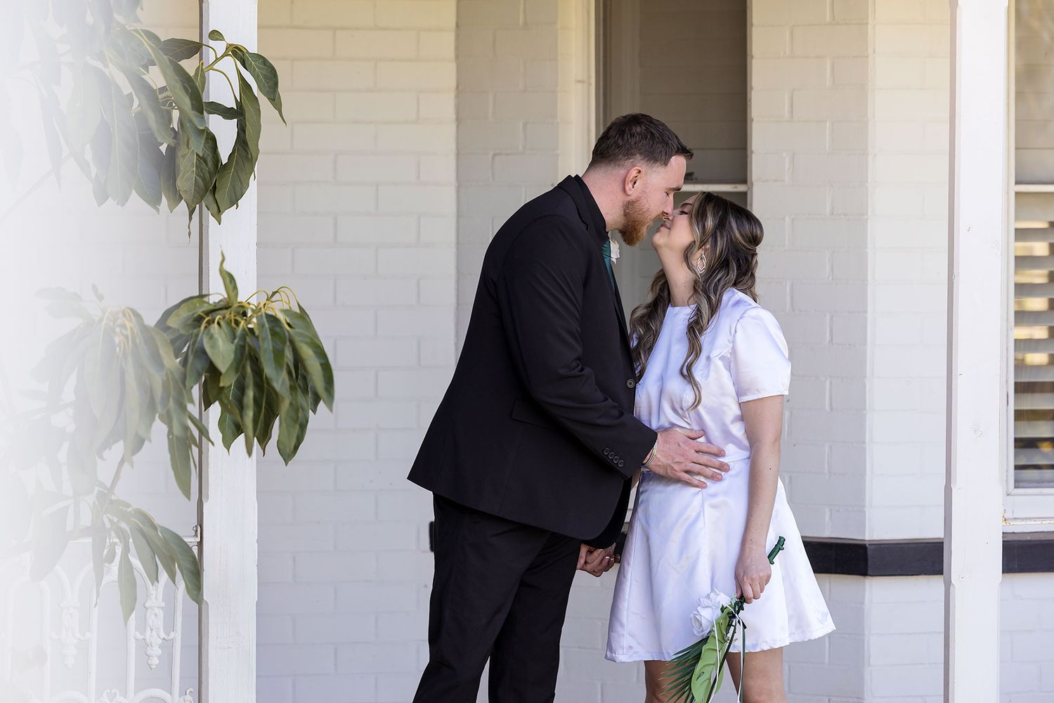 A bride and groom stand together and share a kiss on the porch steps of a classic homestead with white trim at Kimo Estate Gundagai.