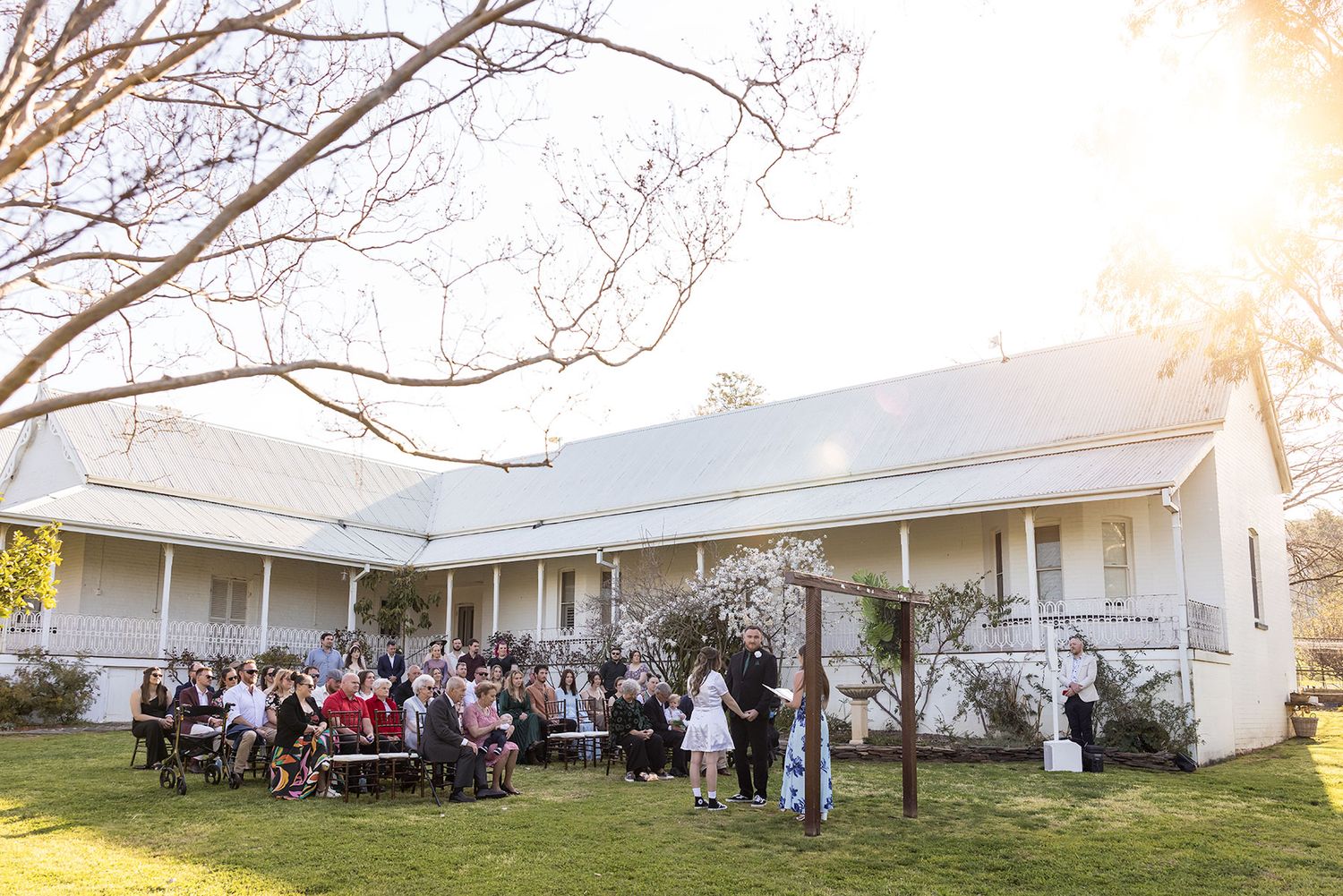 A panoramic view of people gathering outside a single-story white building at sunset with Kimo Estate in the background.