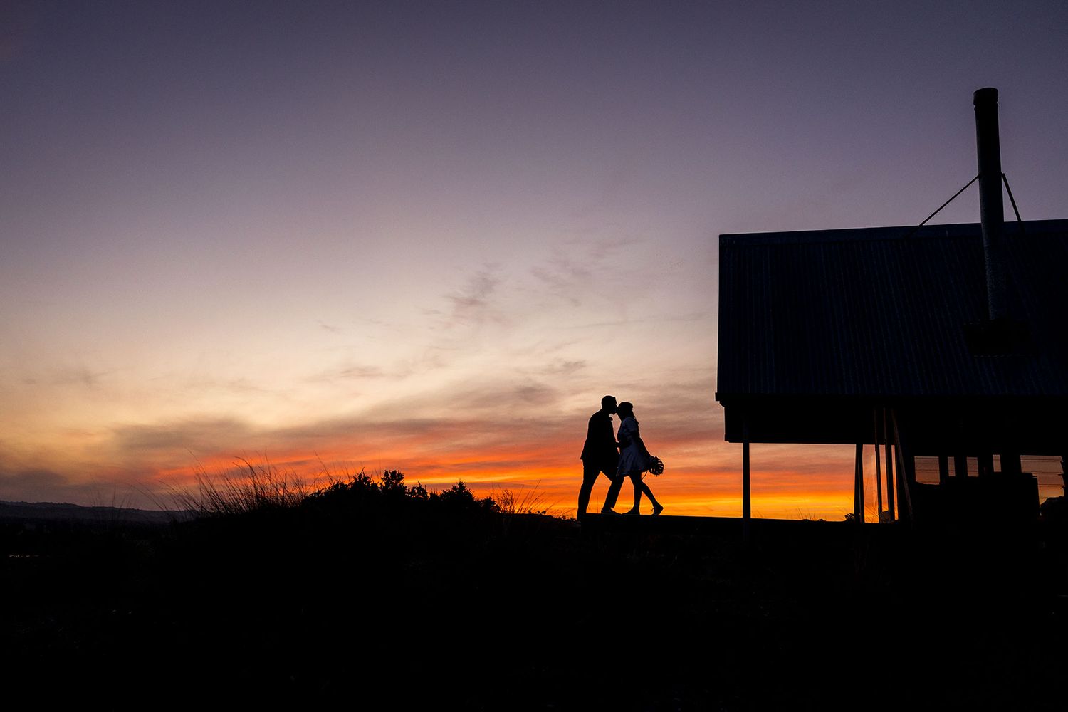 Silhouetted figures of a Bride and Groom set against a vibrant orange and red sunset sky with dark trees on the horizon at Kimo Estate.