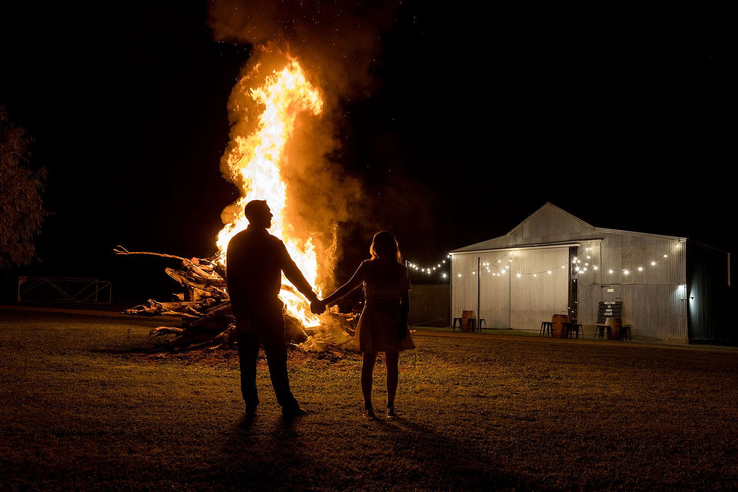 A silhouette of a Bride and Groom holding hands standing in front of a bonfire with the reception venue in the background at Kimo Estate.