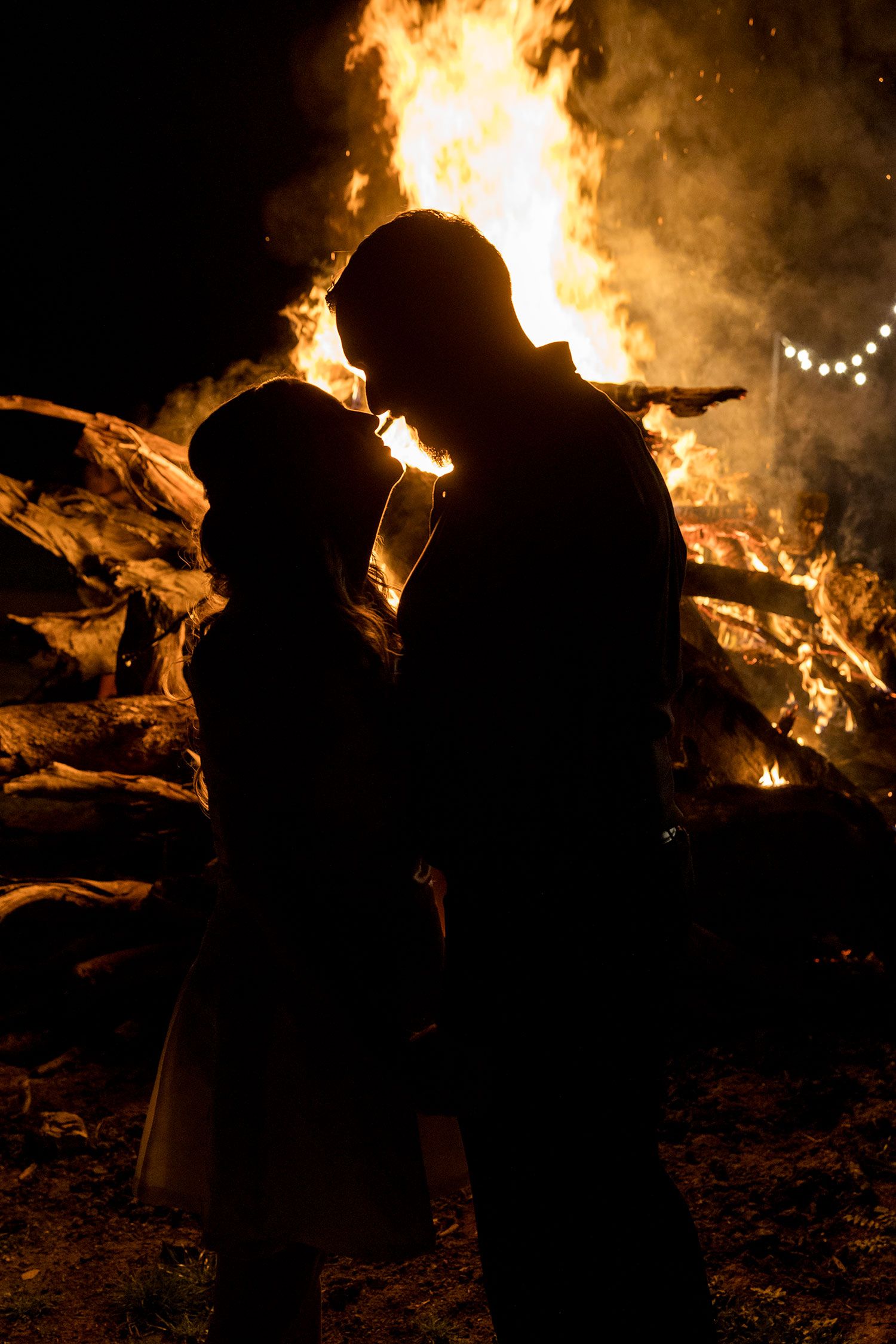 A silhouette of a Bride and Groom holding hands standing in front of a bonfire with the reception venue in the background at Kimo Estate.