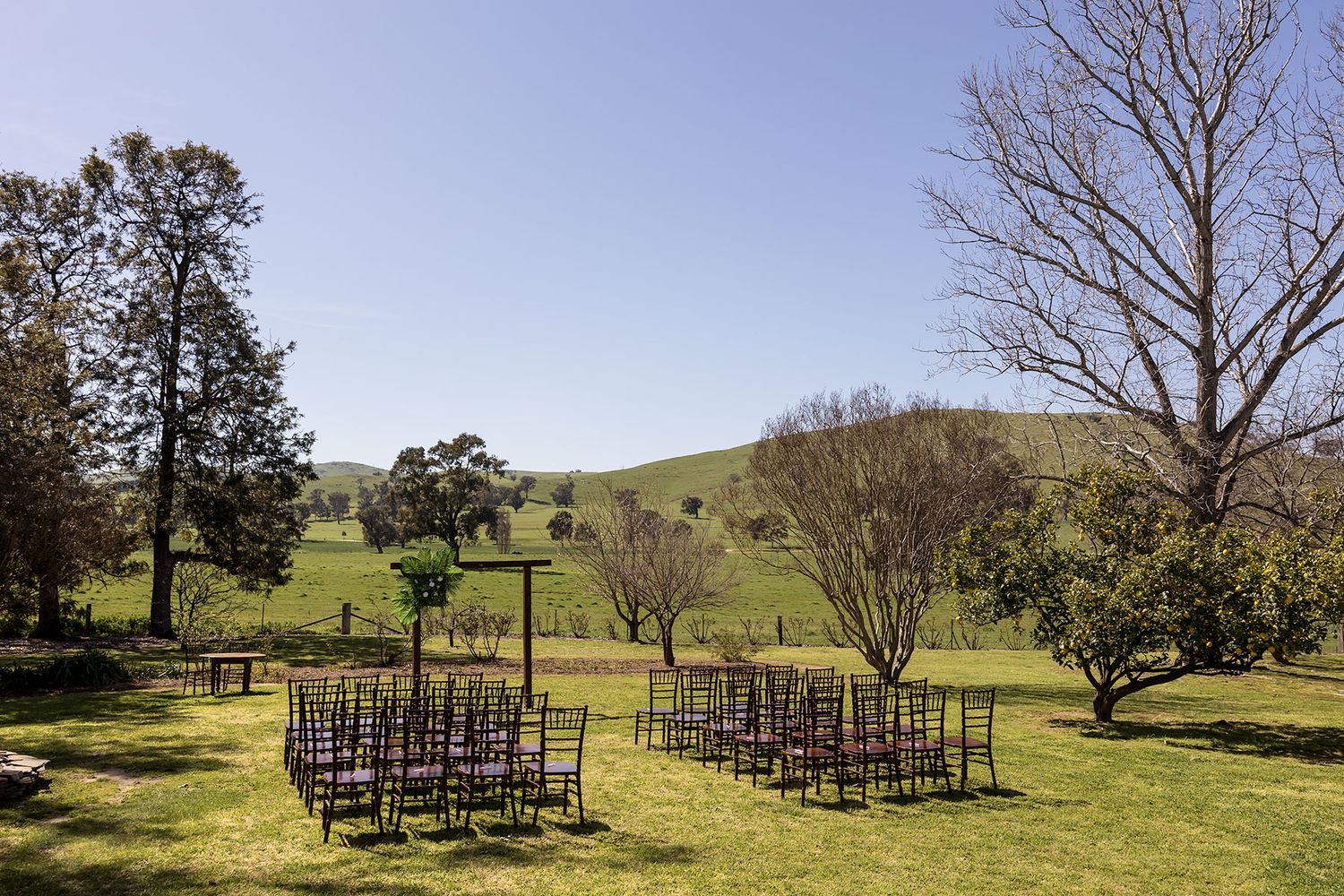 Rows of wooden chairs arranged on a grassy hillside with tall trees and rolling hills in the background ready for a wedding ceremony at Kimo