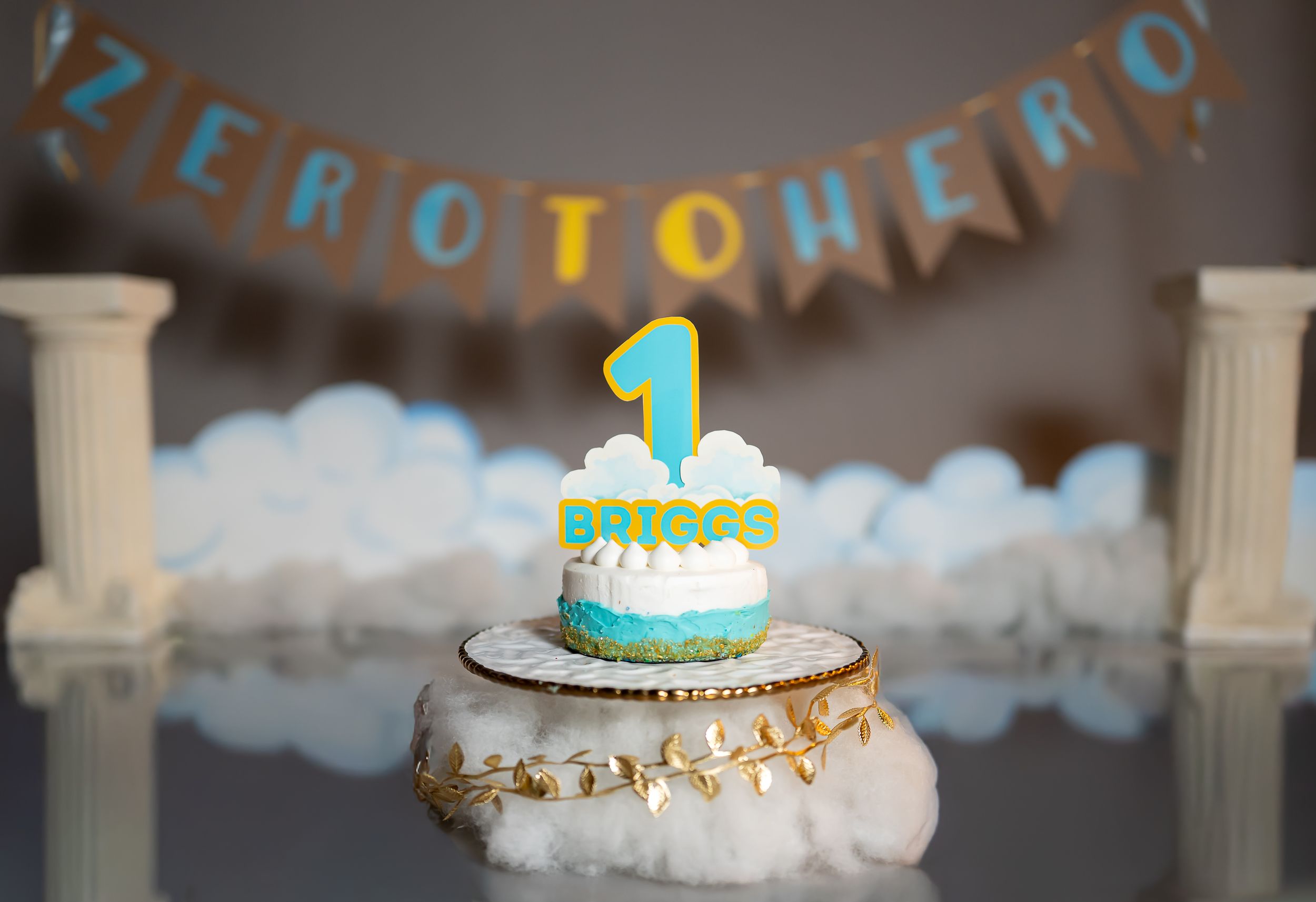 Decorated first birthday cake with blue banner and fluffy white cloud decorations against dark background.
