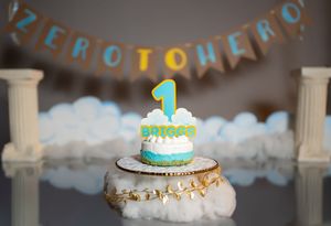 Decorated first birthday cake with blue banner and fluffy white cloud decorations against dark background.