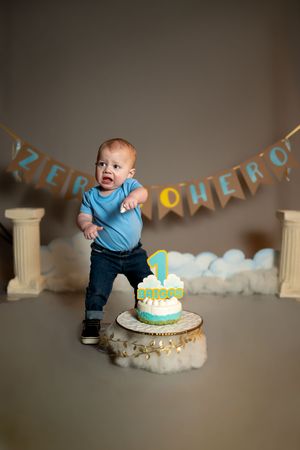 First birthday celebration setup with blue and yellow decorations, cake on a stand, and white pillars against grey backdrop.