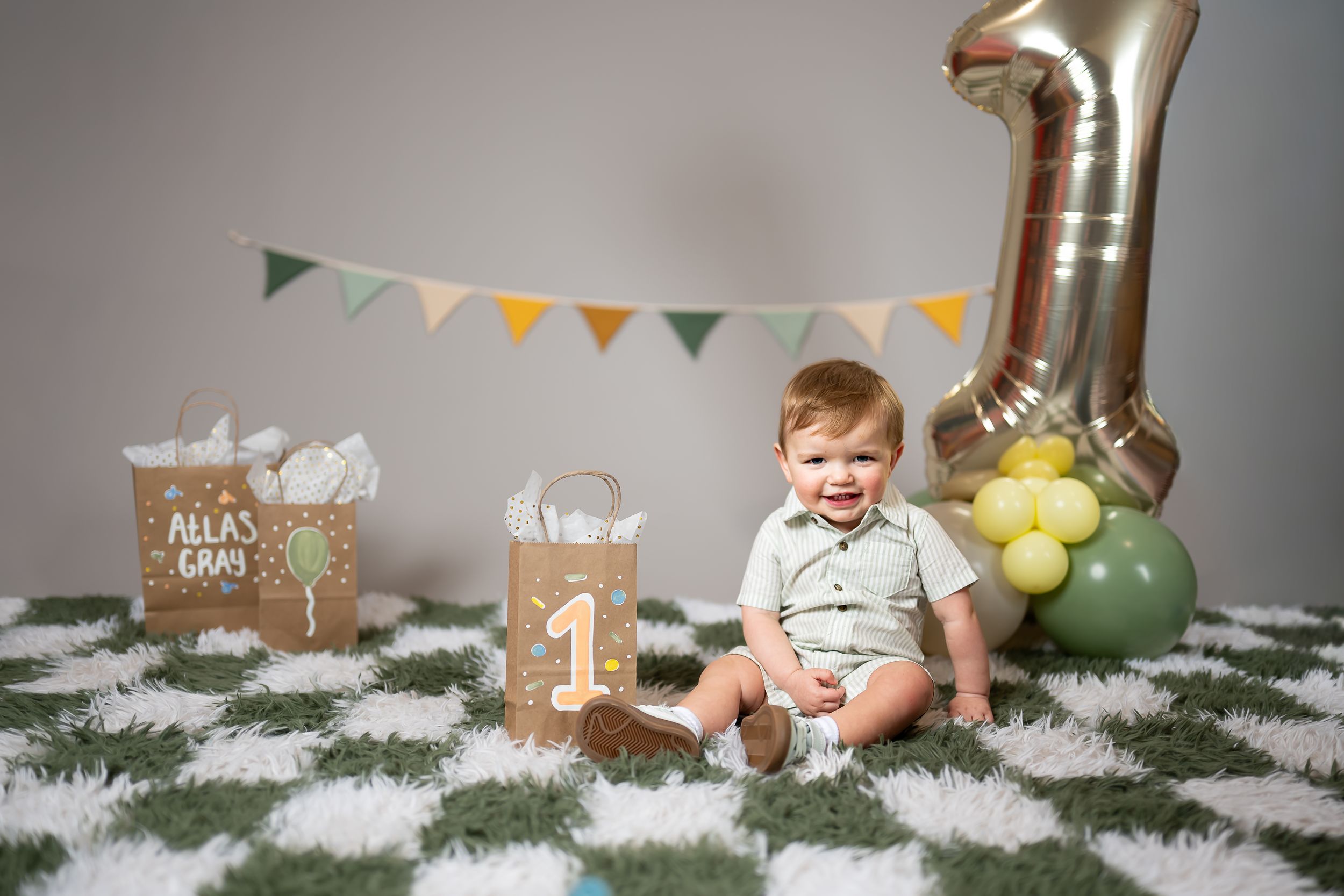 Baby's first birthday cake smash photo with number 1 balloon, bunting, and gift bags on a white fluffy rug.