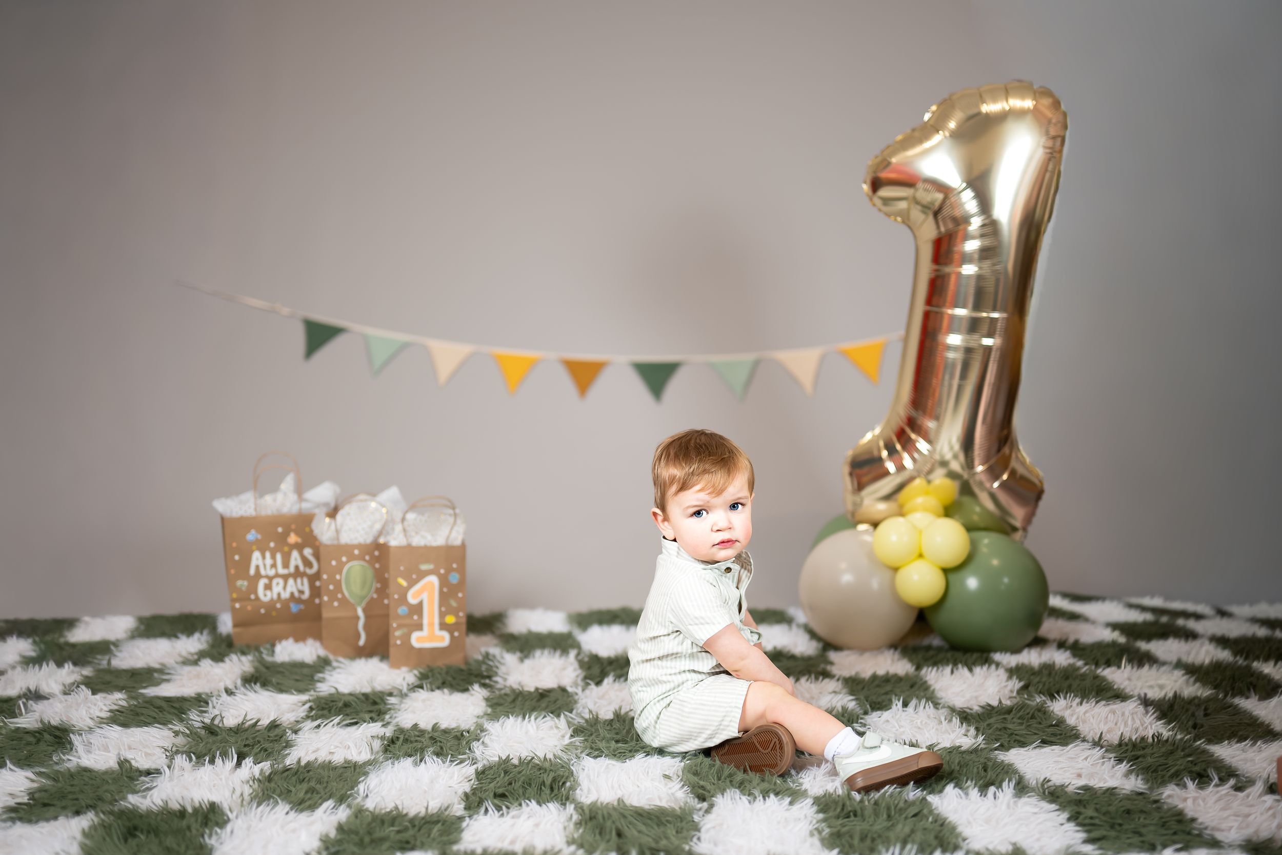 Baby's first birthday cake smash photo with gold number one balloon, bunting, and decorative gift bags on a green rug.