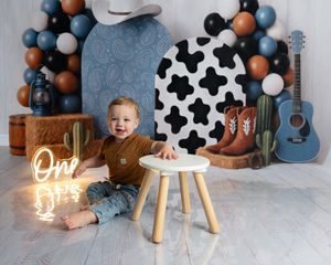 A smiling baby boy sits on a white floor surrounded by a cowboy-themed backdrop with balloons, boots, and a neon “One” sign.