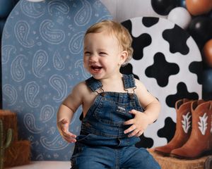 Laughing toddler in denim overalls poses before cow print backdrop at a Western-themed first birthday party.