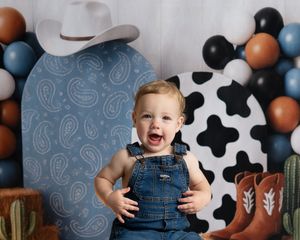 A laughing baby boy in denim overalls sits in front of a cowboy-themed backdrop with cow print and balloon garland.