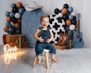 Smiling baby in denim overalls sits on stool at western-themed first birthday party with balloons and guitar.