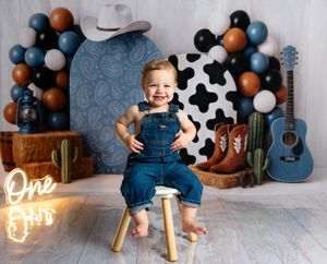 Toddler in denim overalls poses on stool at cowboy-themed first birthday party with balloons and guitar.