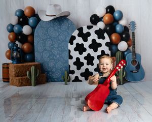 A smiling baby holds a red toy guitar in front of a cowboy-themed backdrop with balloons, cow print, and a blue guitar.