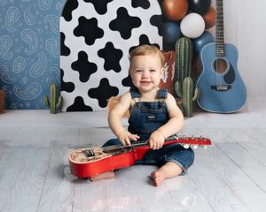 Smiling baby in denim overalls playing with a red toy guitar, with cactus and cow print backdrop.