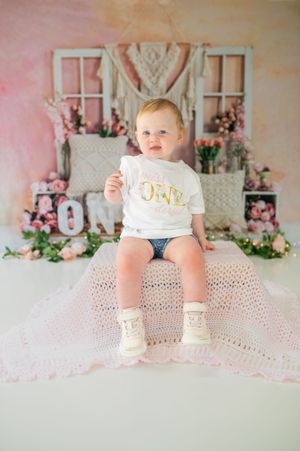 First birthday photoshoot setup with white macrame backdrop, pink florals and a small infant wearing a "ONE" shirt.