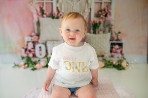 A first birthday photoshoot scene featuring floral decorations, white outfits, and pink and gold party decor.