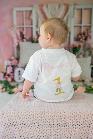 Small child wearing white t-shirts with tropical designs crawling on textured pink surface.
