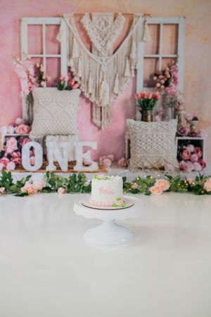 Pink and white first birthday cake setup with macrame wall hanging, floral decorations and letter blocks spelling ONE.