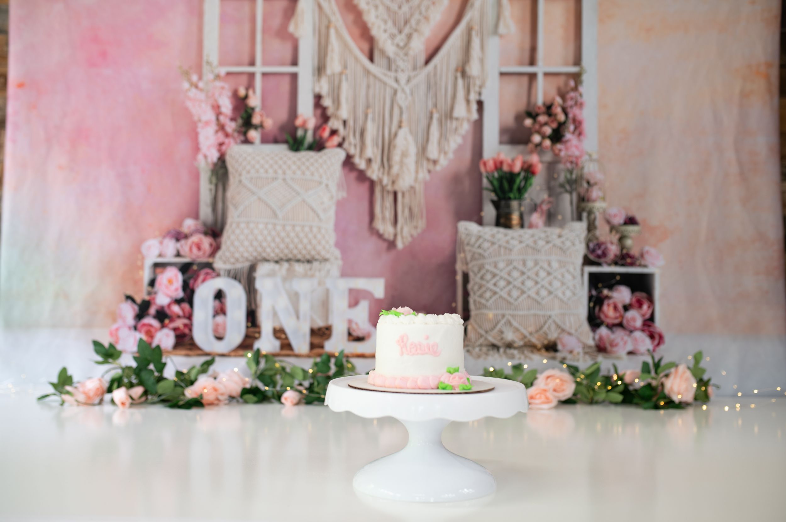 First birthday party setup with macrame backdrop, pink flowers, 'ONE' letters, and white cake on pedestal stand.