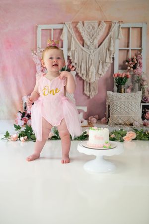 First birthday photo setup with pink tulle outfit, white cake stand, and boho macrame wall decoration.
