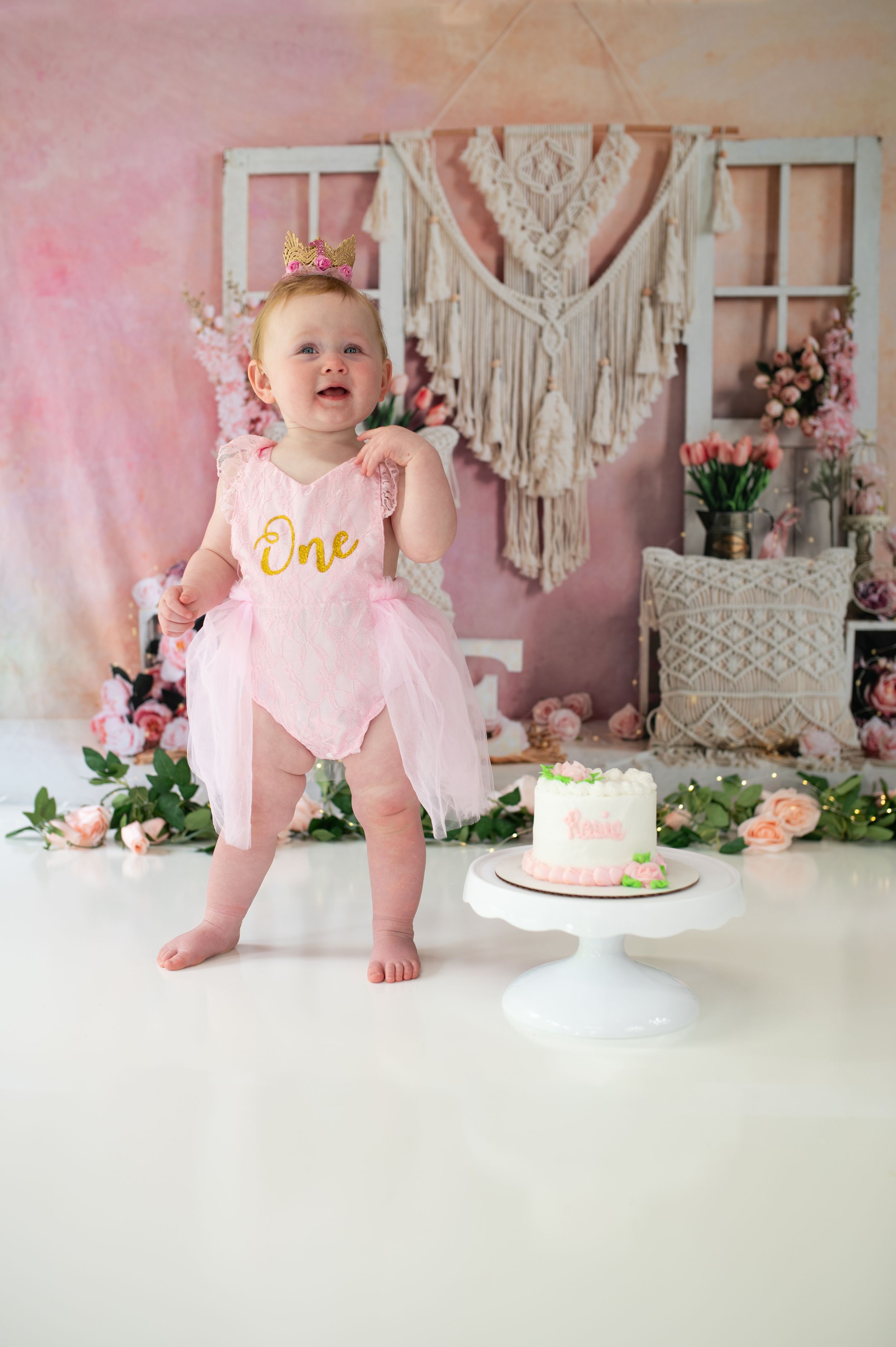 First birthday photo setup with pink tulle outfit, white cake stand, and boho macrame wall decoration.