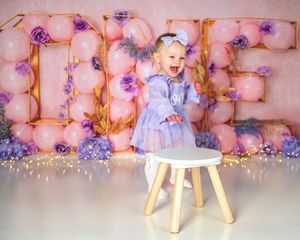 A small wooden stool sits against a pink floral wall with purple accents and string lights creating a romantic atmosphere.
