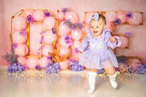 Illuminated letters spelling ONE with purple flowers create a charming backdrop for first birthday photoshoot setup.
