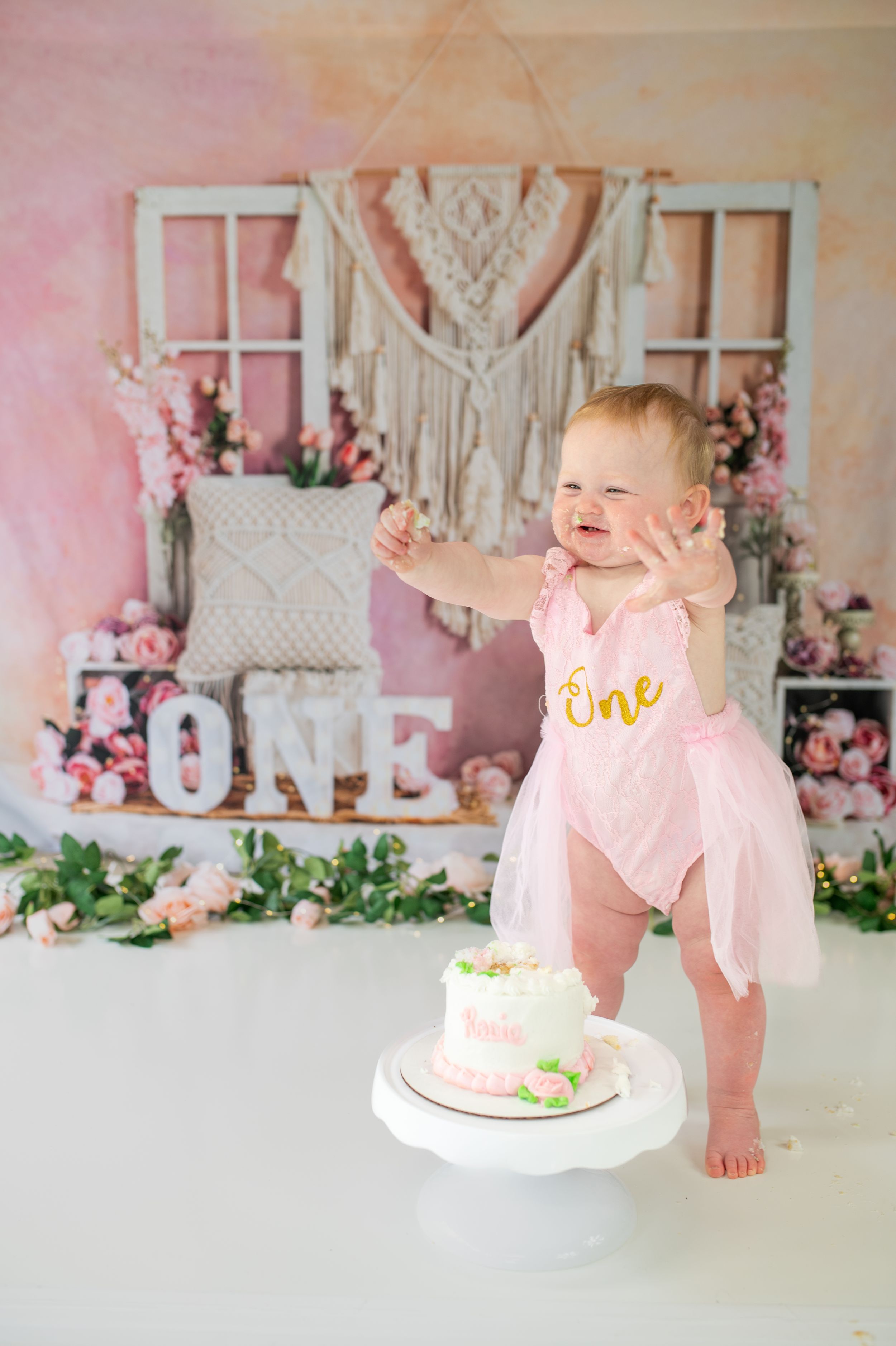 Pink and white vintage first birthday cake smash photo session setup with floral decorations and rustic decor elements.
