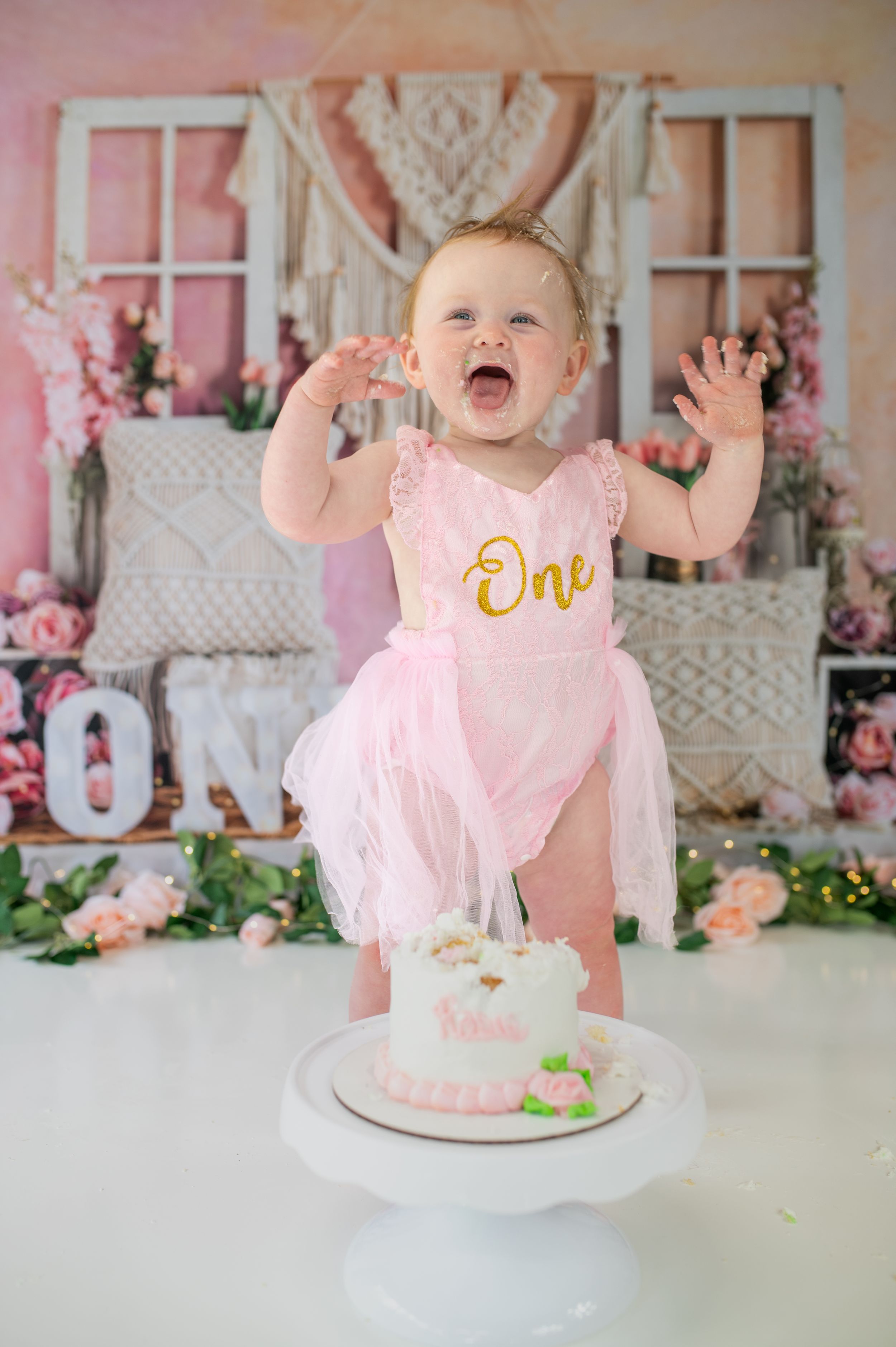 Baby in pink tutu dress celebrating first birthday with cake smash photoshoot against floral backdrop.