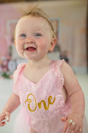 Baby in pink lace dress smiles while wearing cake smudges during first birthday cake smash photo session.
