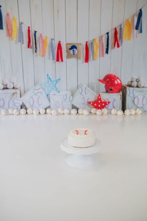 White cake on pedestal with red toadstool cake topper against backdrop of colorful fabric bunting and white decor.
