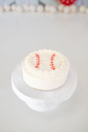 White frosted cake with baseball stitching design sits on a simple white cake stand against blurred background.