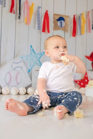 Baby wearing blue pants and white shirt enjoys smashing and playing with cake at a baseball themed party setup.