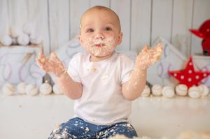 A baby wearing a white shirt smiles while playing with and getting messy with colorful cake frosting during a special celebration.
