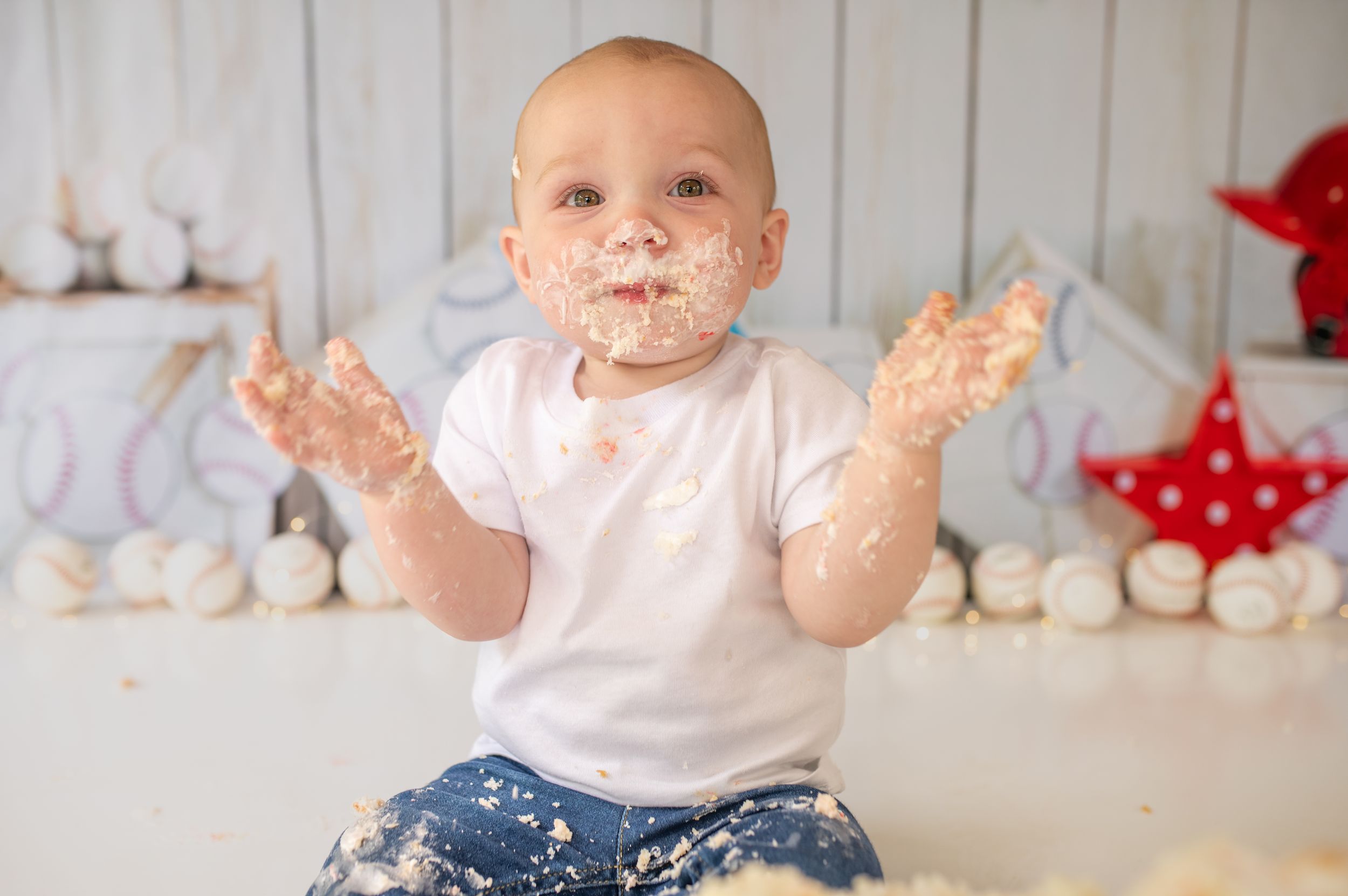 A baby wearing a white shirt smiles while playing with and getting messy with colorful cake frosting during a special celebration.