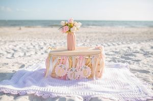Pink and white themed baby announcement display with flowers and banners set up on a white blanket at the beach.
