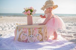 Baby celebration display with pink decorations, floral arrangements, and bunting on a blanket at a sunny beach.