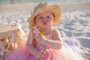 A child wearing a straw hat enjoys sitting and playing in the white sand on a sunny beach.