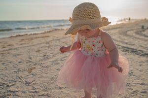 Baby wearing a pink tutu dress and straw hat explores sandy beach at sunset during golden hour.